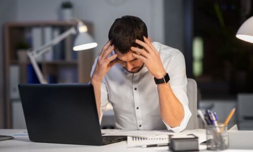 Man at desk in front of laptop, hands on head looking frustrated and defeated.