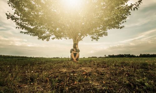 Woman sitting at base of tree with sunshine coming through in an open space contemplating