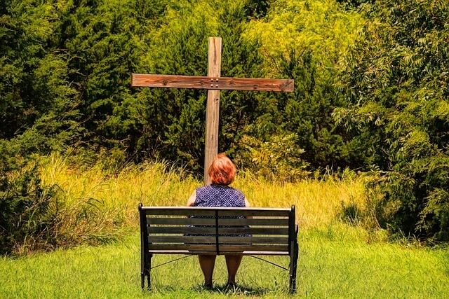 Woman on outside bench in front of wooden cross.