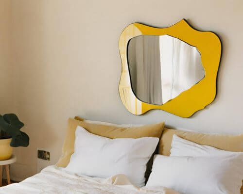 Fun asymmetrical mirror on yellow framing board hung over a bed
